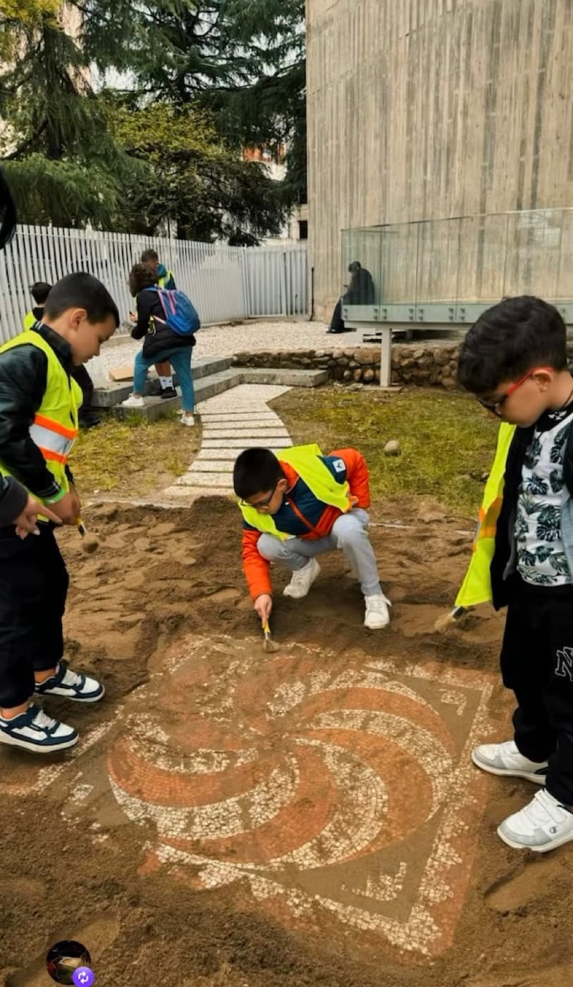 Children in high-visibility vests brushing away sand to reveal a mosaic during a Little Archaeologist workshop at the site.