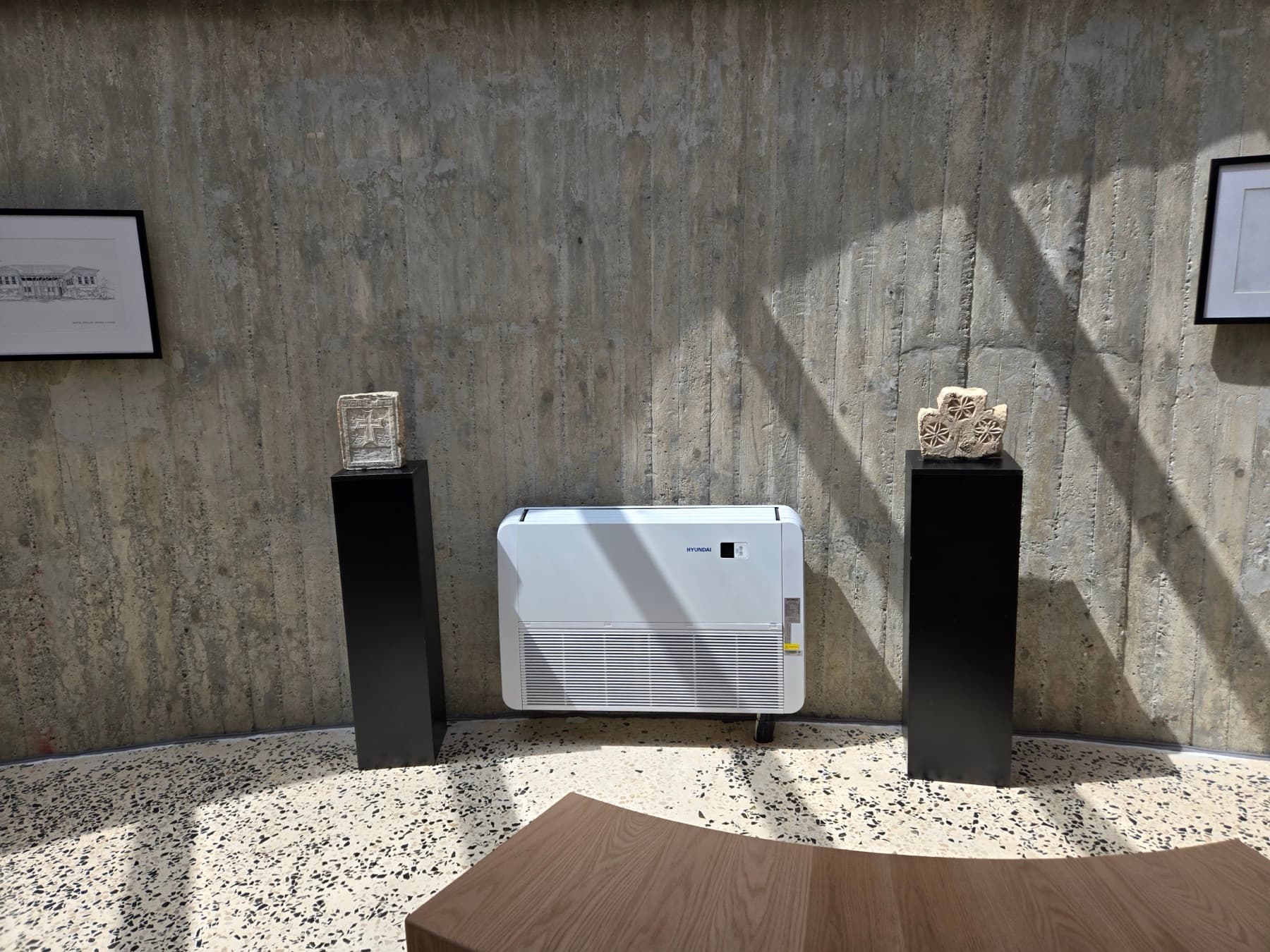 Museum wall with framed prints and two stone artefacts mounted on black pedestals against a raw concrete wall.